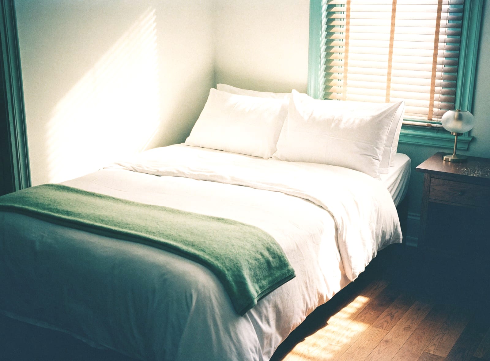 Neatly made bed with white linen and a green throw by a window